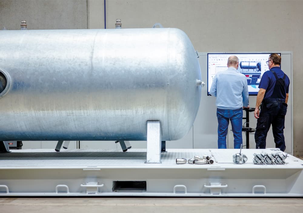 Grosser BOGE Druckluftbehälter auf grauem Containergrund mit zwei BOGE Mitarbeitern am Computer im Hintergrund
