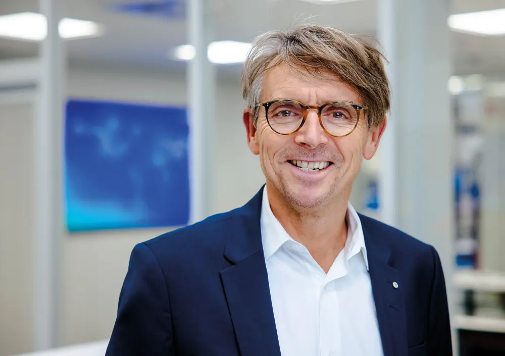 BOGE Marketing Mitarbeiter in dunkelblauem Anzug, Brille und weißem Hemd in einem Buero laechelt in die Kamera