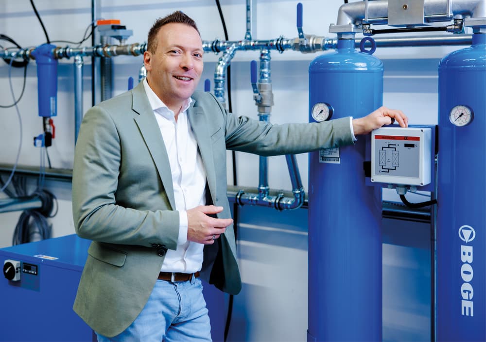 BOGE Druckluftaufbereitungs-Berater in einem grauen Blazer steht lächelnd nebenAufbereitungsanlagen und blauen Behältern und hält ein digitales Gerät in der Hand, in einer sauberen Fabrikumgebung.