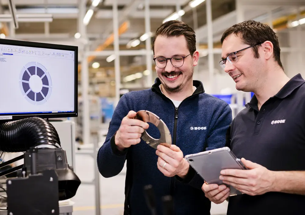 Zwei Männer in einer Fabrik betrachten gemeinsam ein Kompressorbauteil neben einem Computerbildschirm