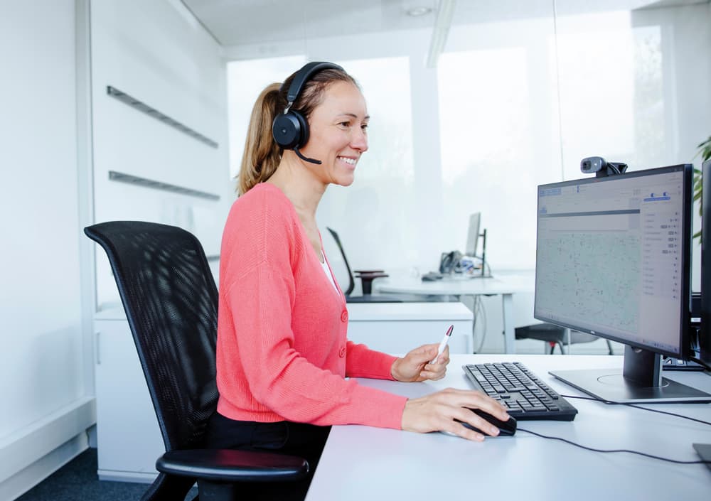 BOGE Kundensupport Mitarbeiterin in koralle-farbenem Pullover mit Headset lächelnd vor einem Computer mit geöffneter Landkarte.