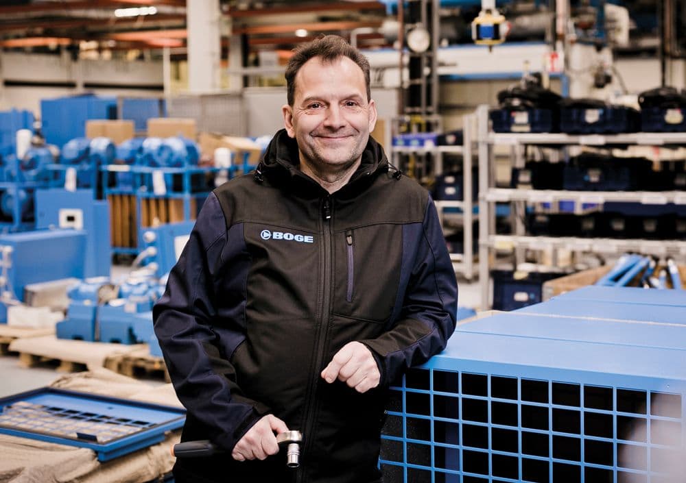 BOGE Monteur an Schraubenkompressor gelehnt mit einem Werkzeug in der Hand in einer Lagerhalle voller Kompressoren und Bauteile.