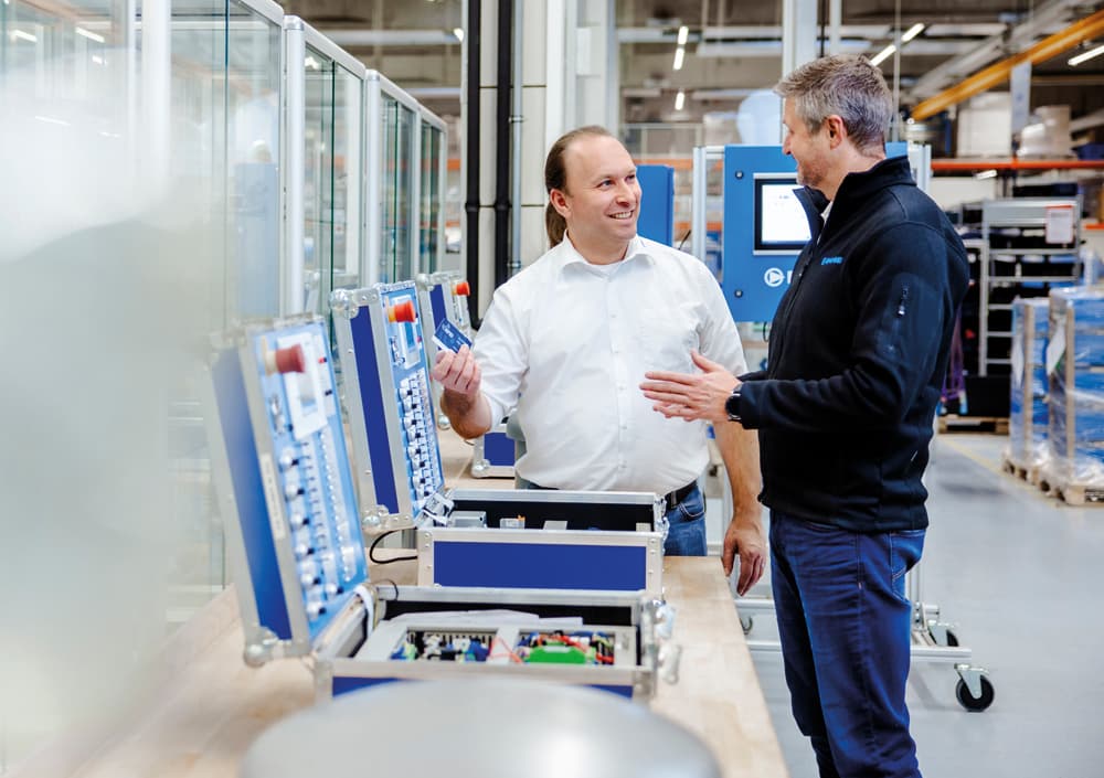 Zwei BOGE Mitarbeiter in Schulungssituation am Steuerungskoffer in einer Werkshalle mit Kompressorsteuerung im Hintergrund