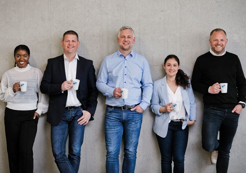 BOGE Mitarbeiter stehe zu fünft vor einer Wand und lehnen sich dagegen, jeder hält eine BOGE Tasse in der Hand und alle lächeln