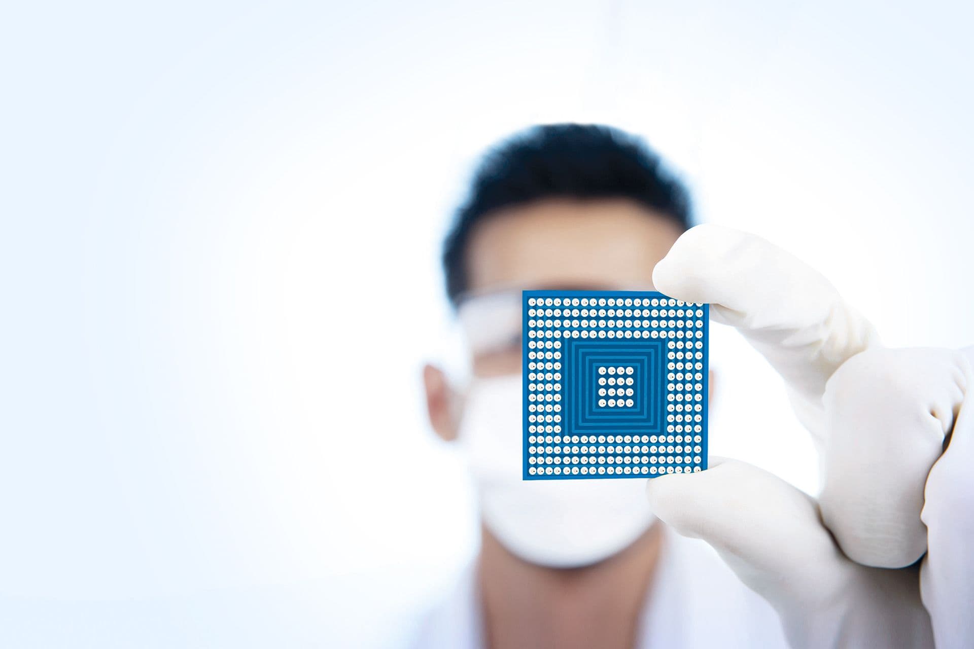 Eine Person mit Handschuhen und Maske hält einen kleinen Computerchip nahe an die Kamera, der Hintergrund ist unscharf.