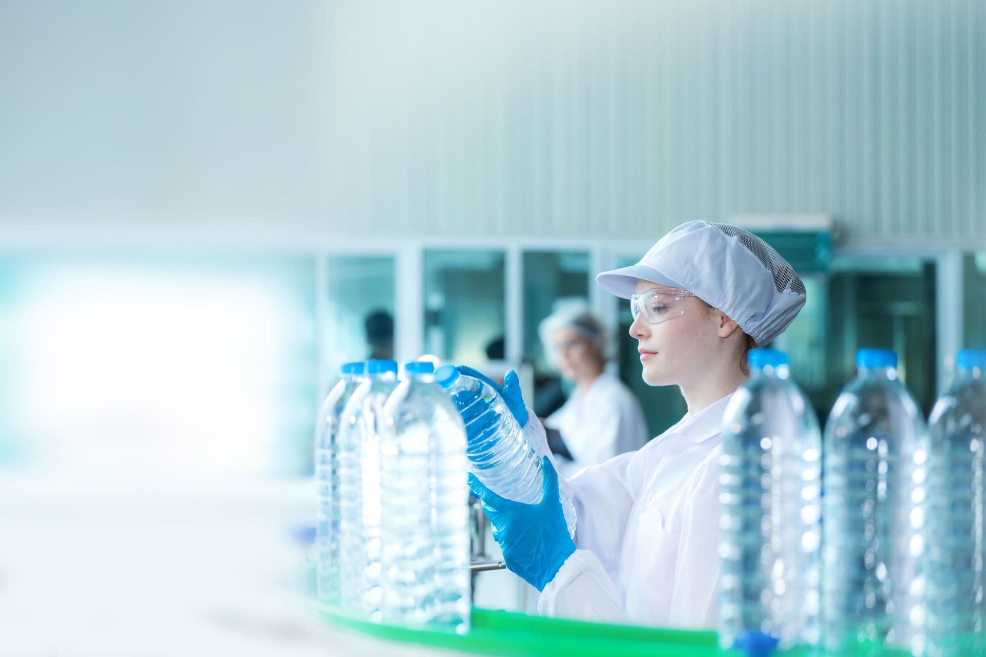 Eine Frau in weißem Arbeitsanzug begutachtet Flaschen auf einem Getränkeabfüllband.