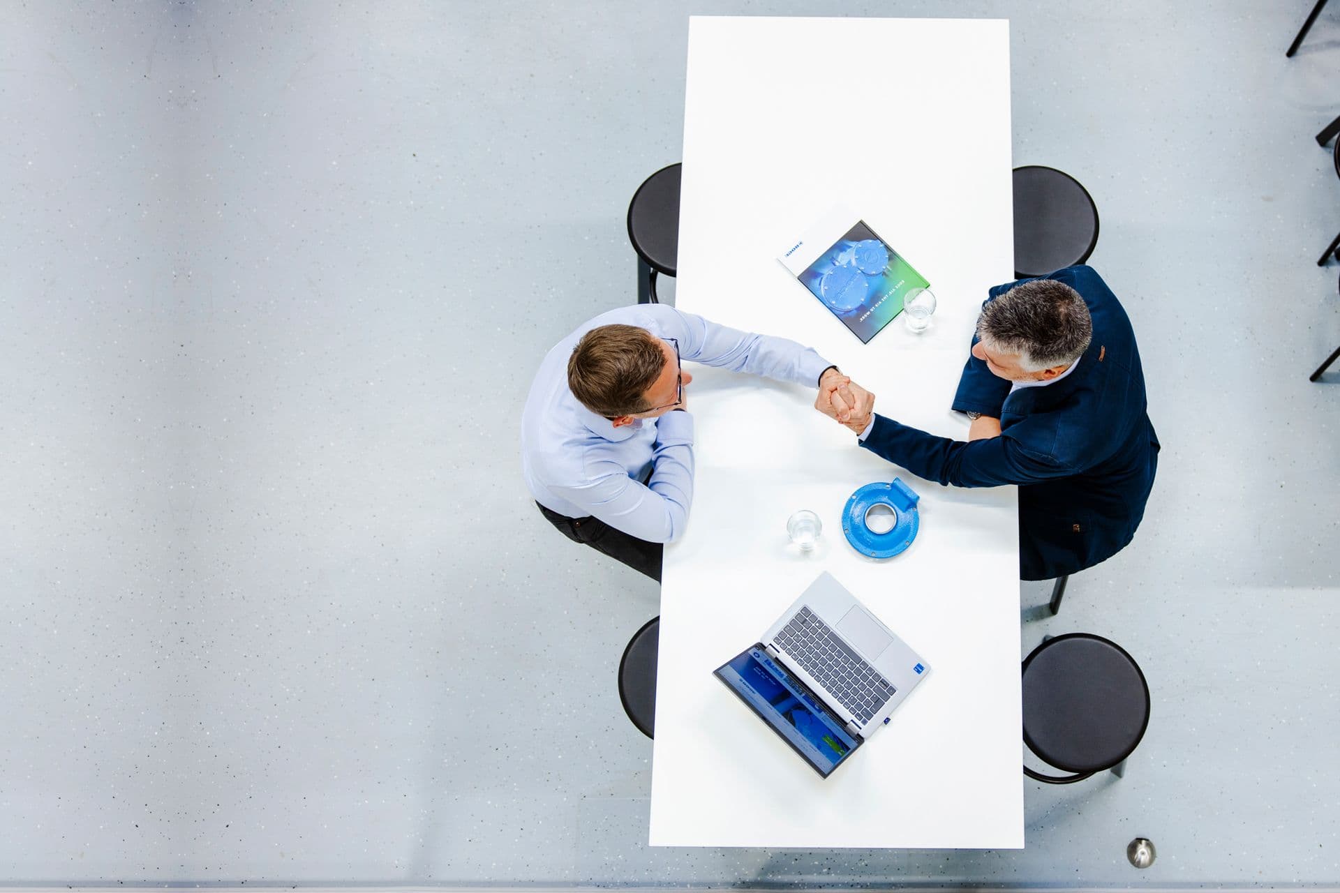 Two people shaking hands across a white table with a laptop, documents, and cups. Viewed from above in a modern office setting.