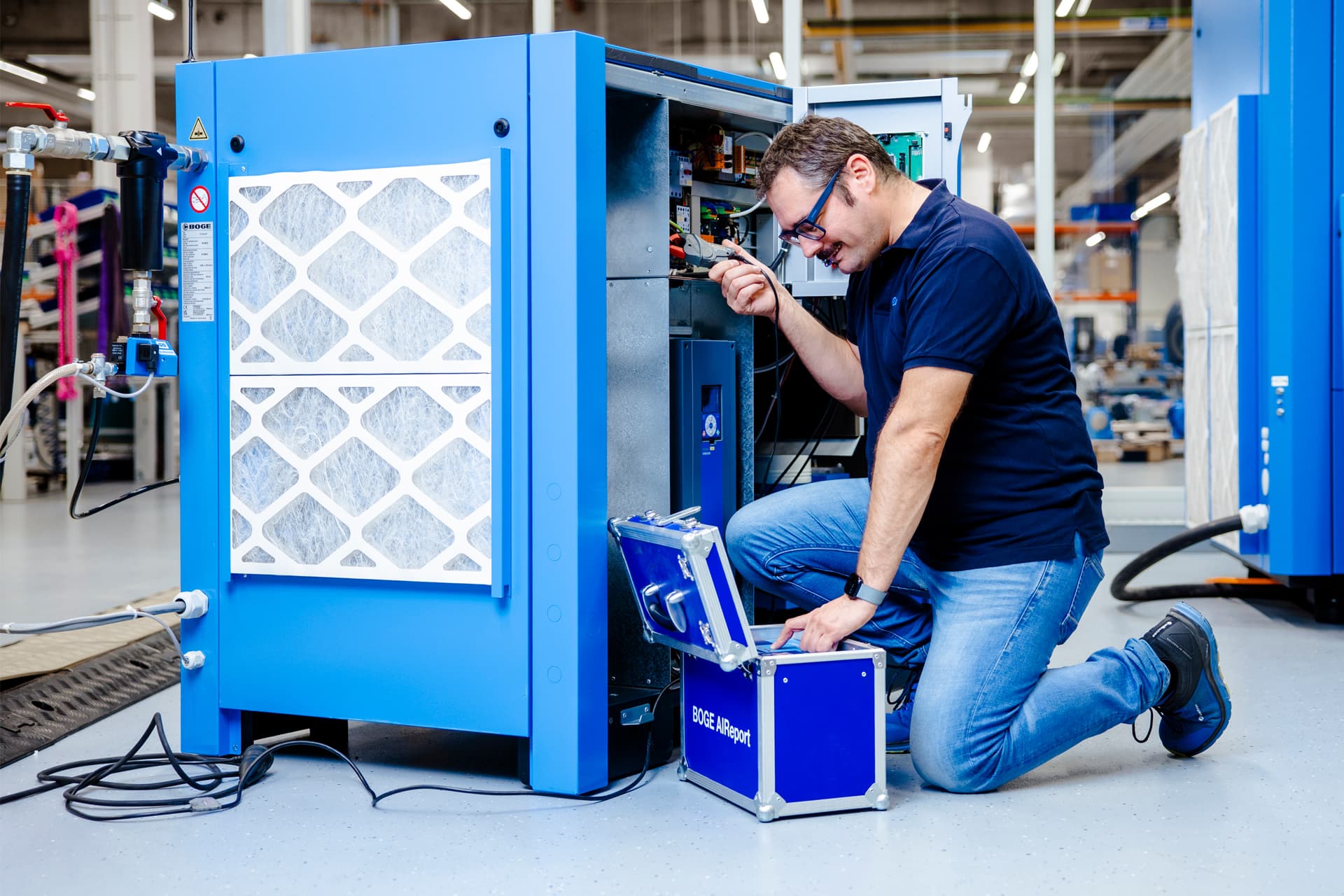 BOGE Servicetechniker fuehrt laechelnd eine Druckluftmessung durch, waehrend er kniend neben dem Kompressor sein Messgeraet bedient.