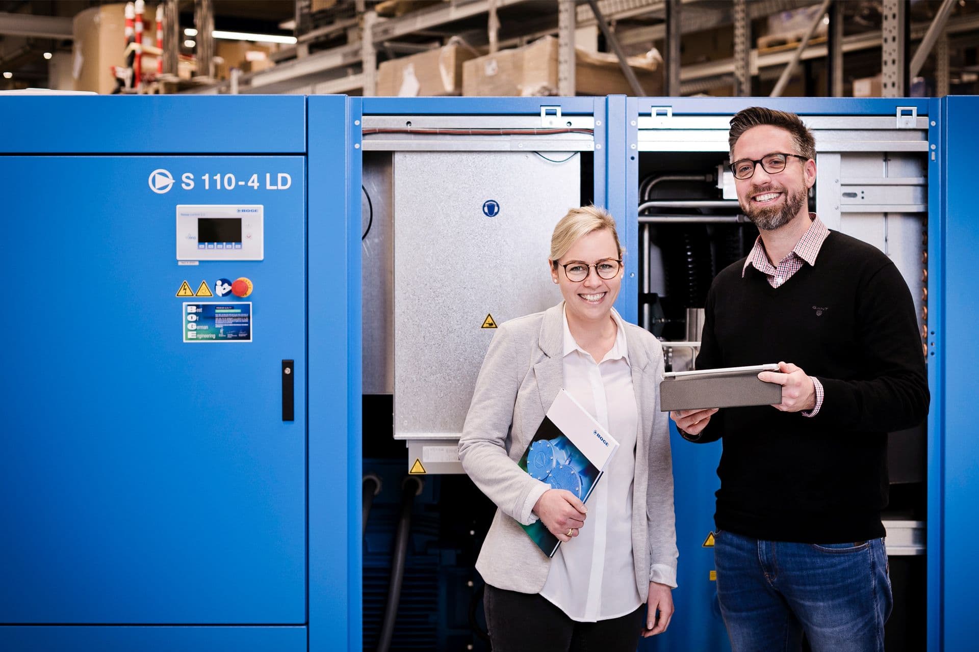 Zwei BOGE Mitarbeiter stehen laechelnd vor einem Schraubenkompressor, genau genommen vor einer S 110-4 LD