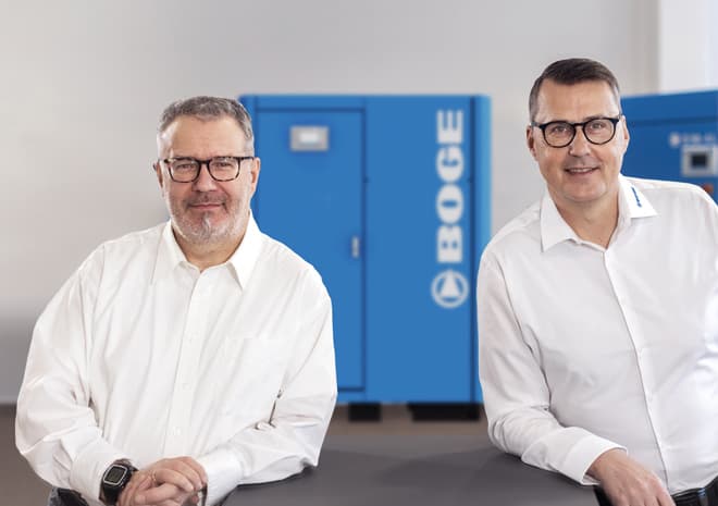 BOGE Gebraucht- und Mietkompressoren Ansprechpartner lehnen sich auf einen Tisch, im Hintergrund ist ein BOGE Kompressor zu sehen.